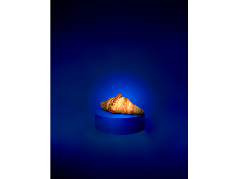 A golden croissant on a blue pedestal against a dark blue background.