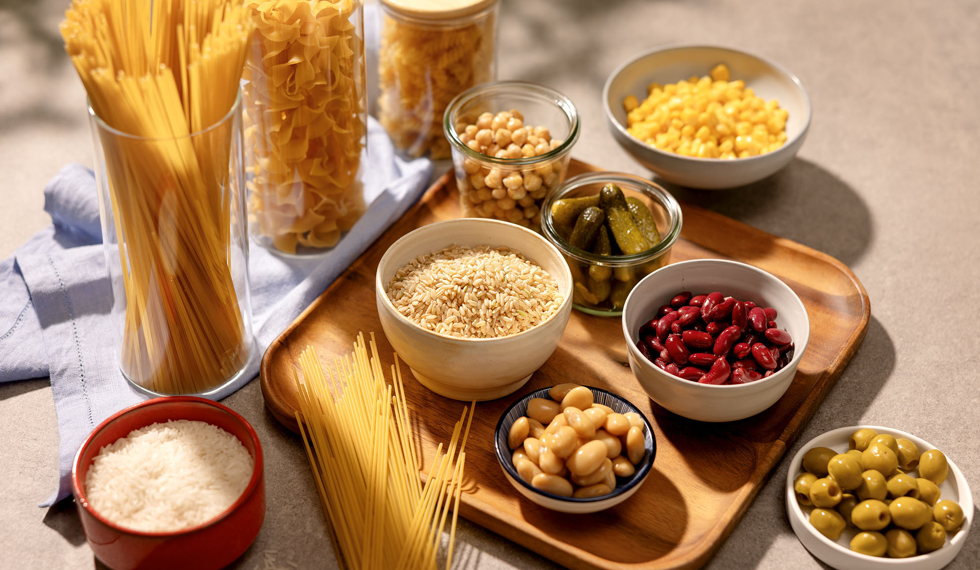 Assortment of food items: spaghetti, pasta, rice, corn, beans, chickpeas, pickles, and olives.