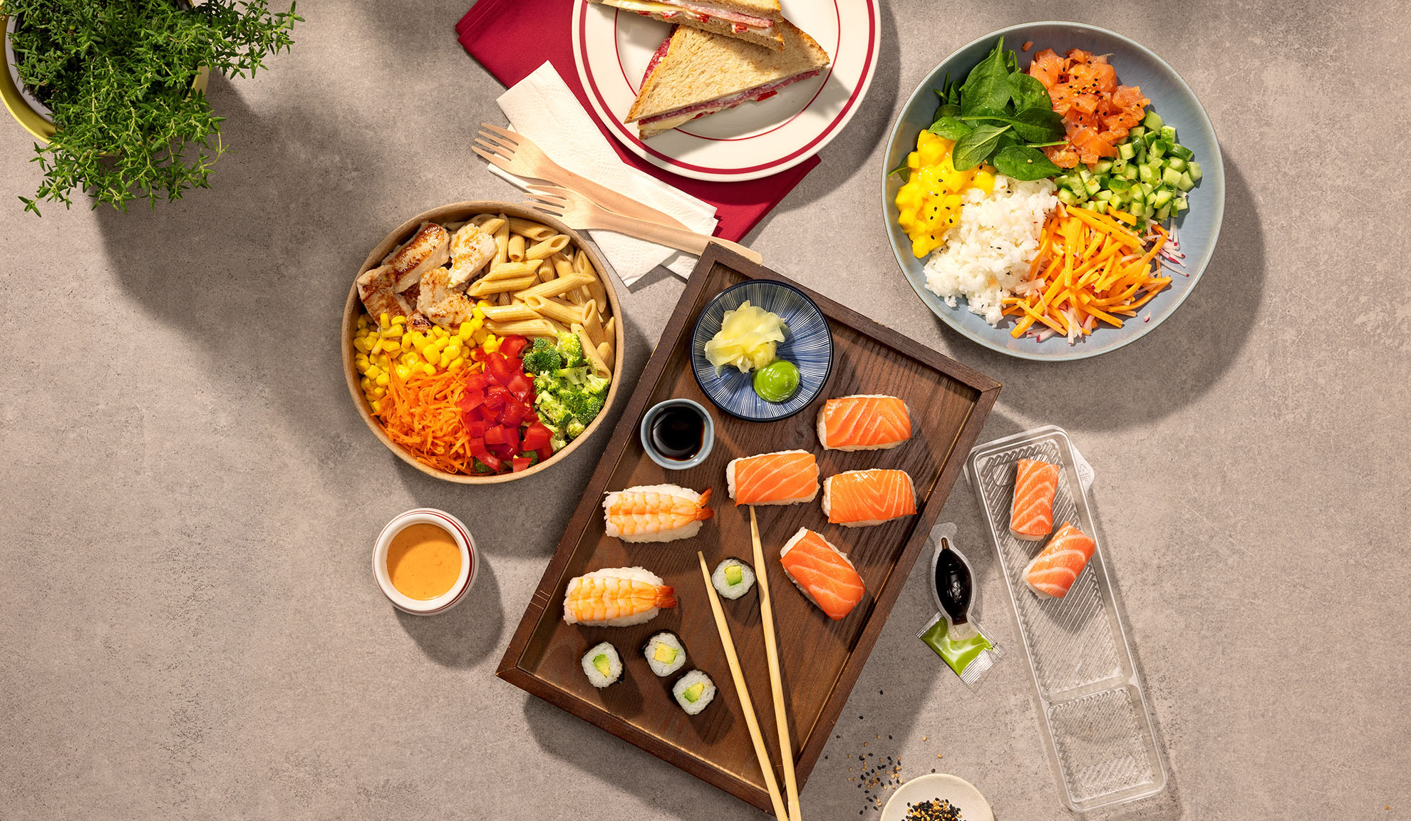 Overhead shot of various takeaway meals: sushi, poke bowl, pasta salad, and sandwiches.
