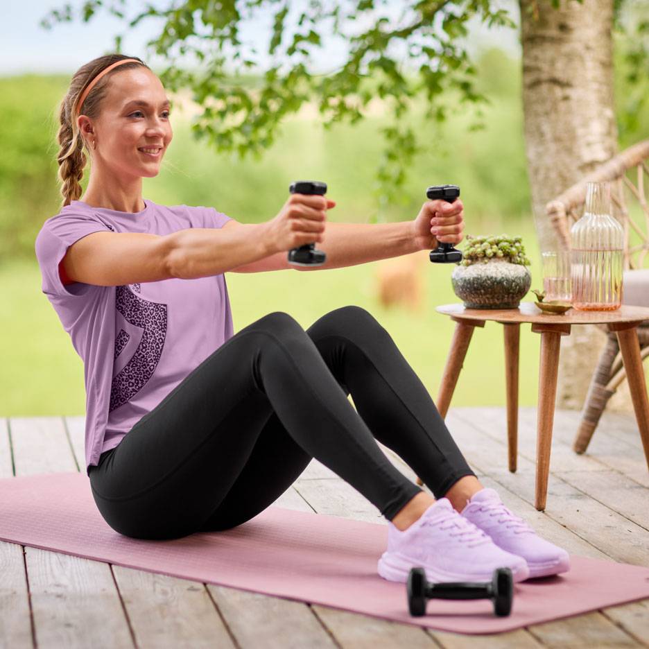 Woman in purple t-shirt and black leggings exercising with dumbbells on a yoga mat.