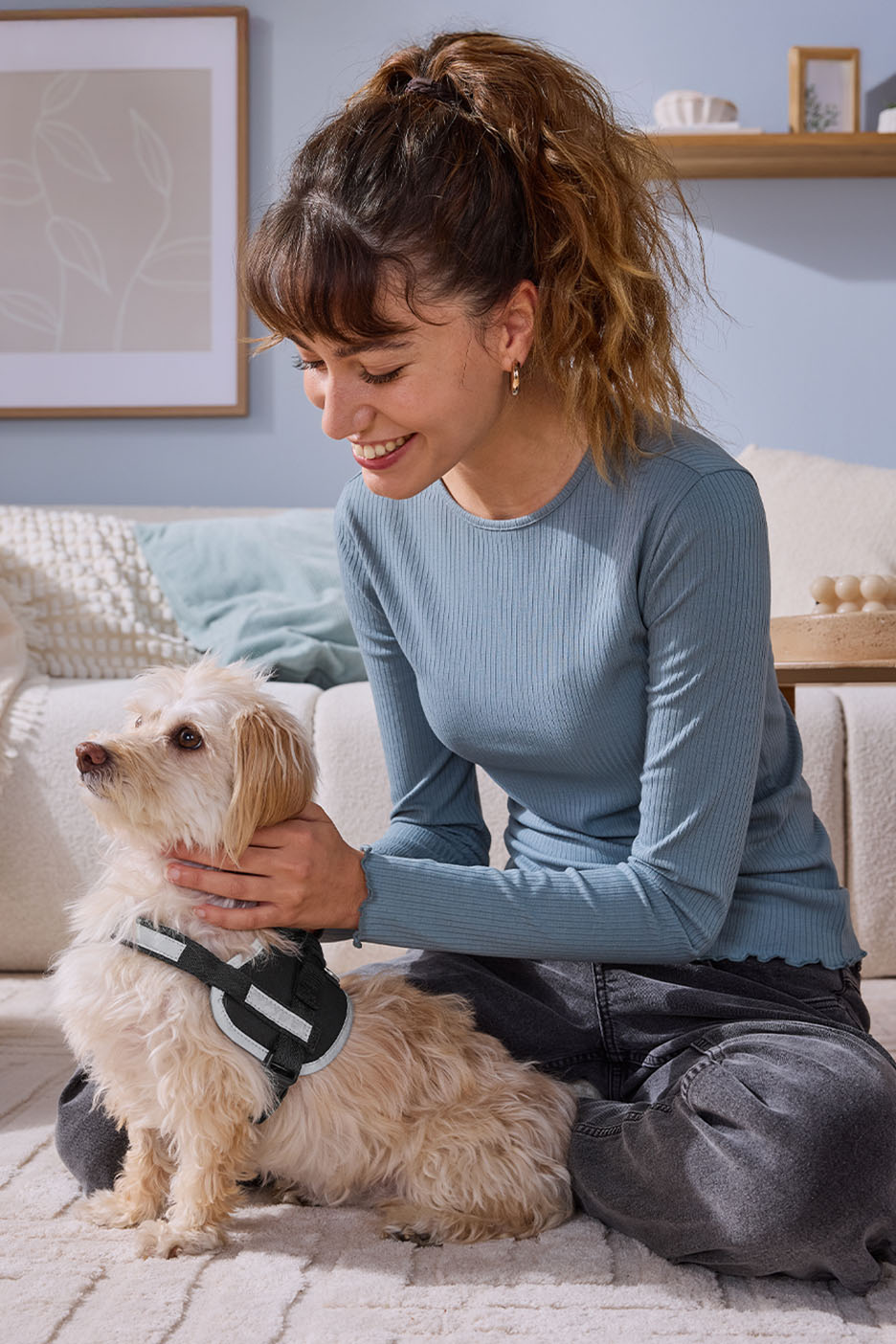 Woman in blue ribbed top and dark jeans petting a small dog with a harness.