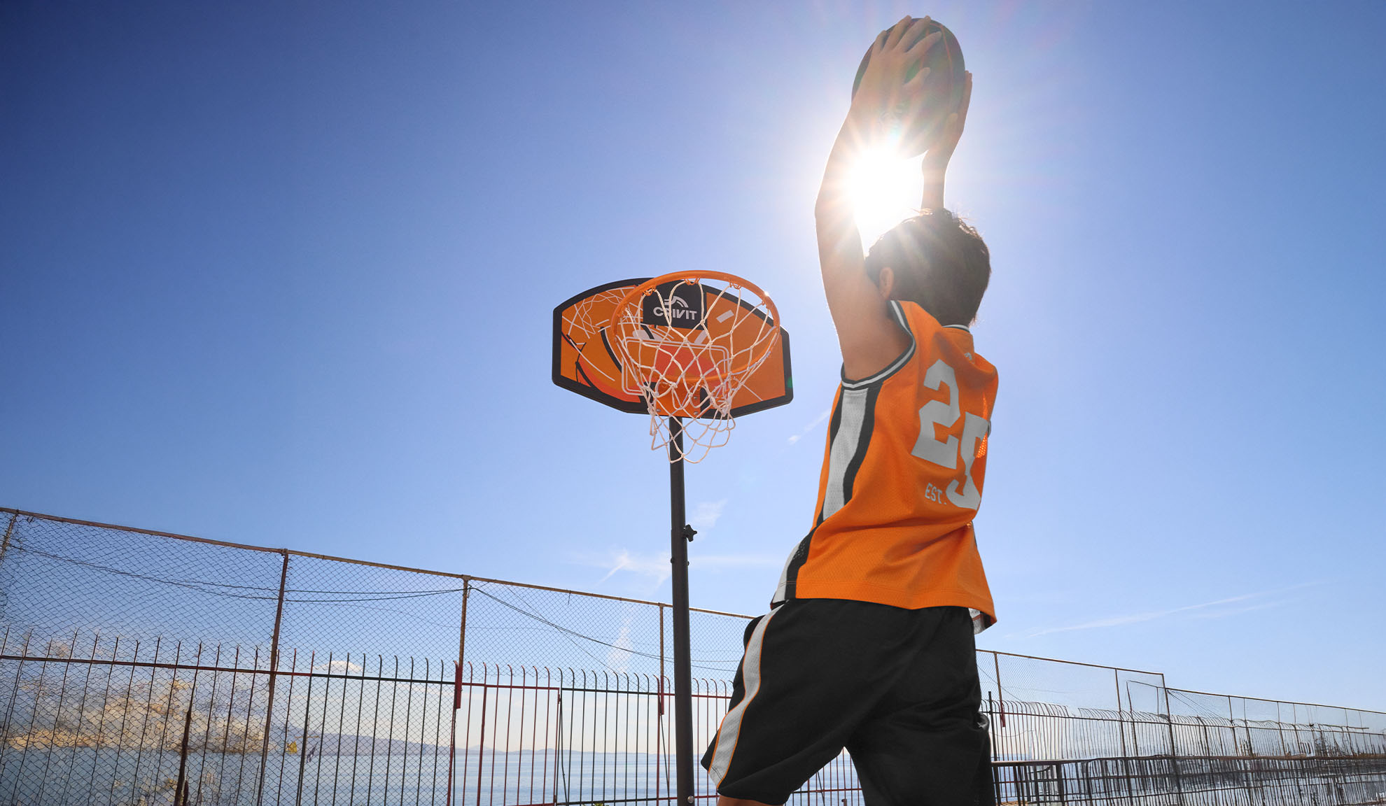 Boy in basketball jersey shooting hoops outdoors under a bright sun.