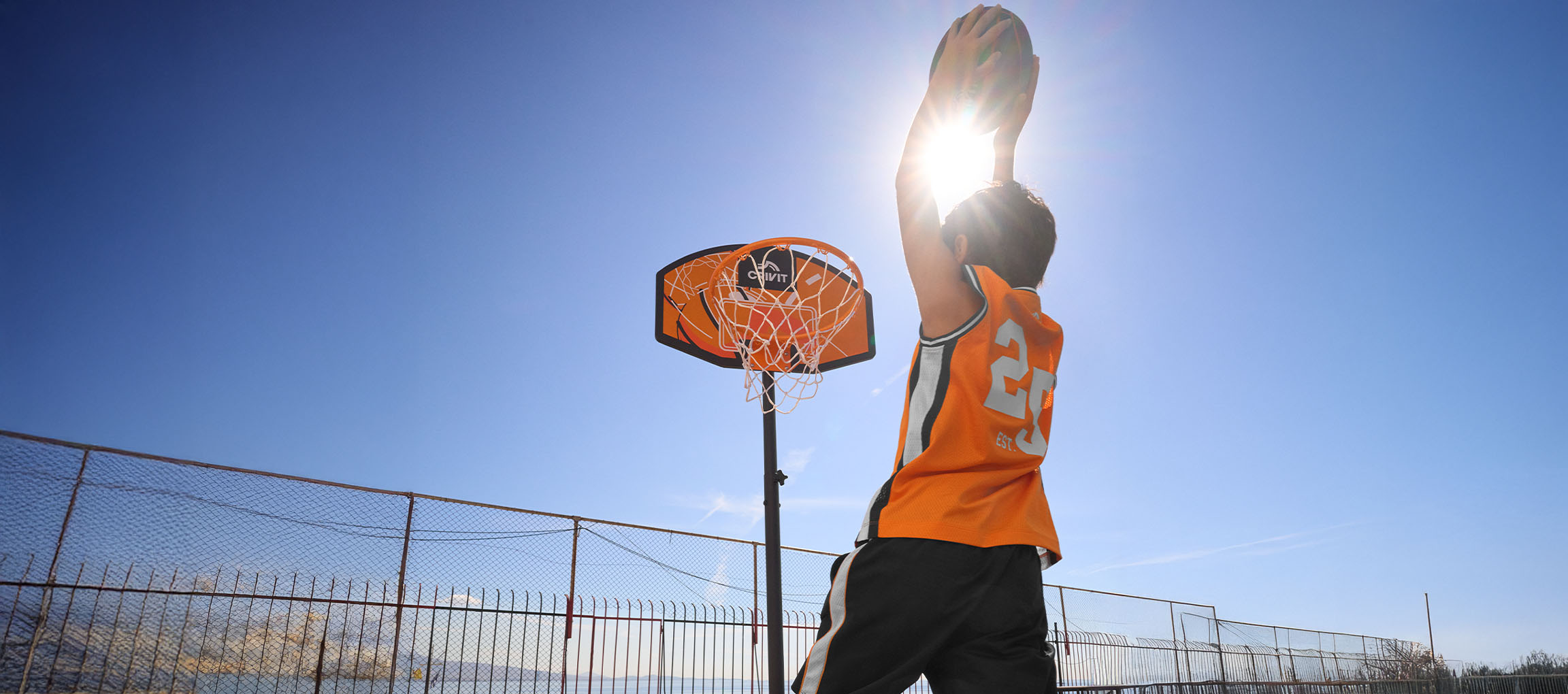 Boy in orange basketball jersey shooting a ball into the hoop on a sunny day.