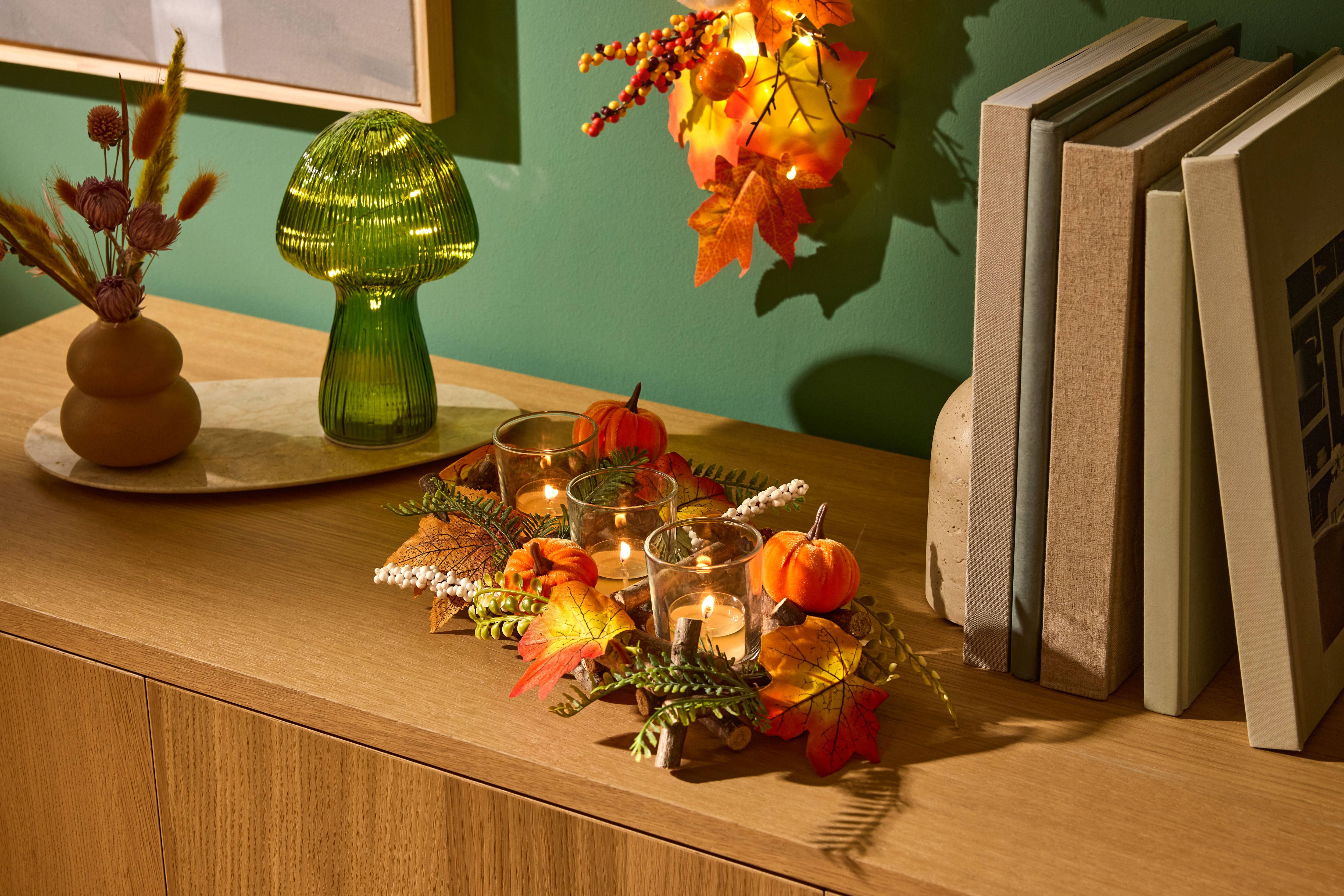 Autumnal table decoration with pumpkins, candles, and leaves, next to a green mushroom lamp and books.