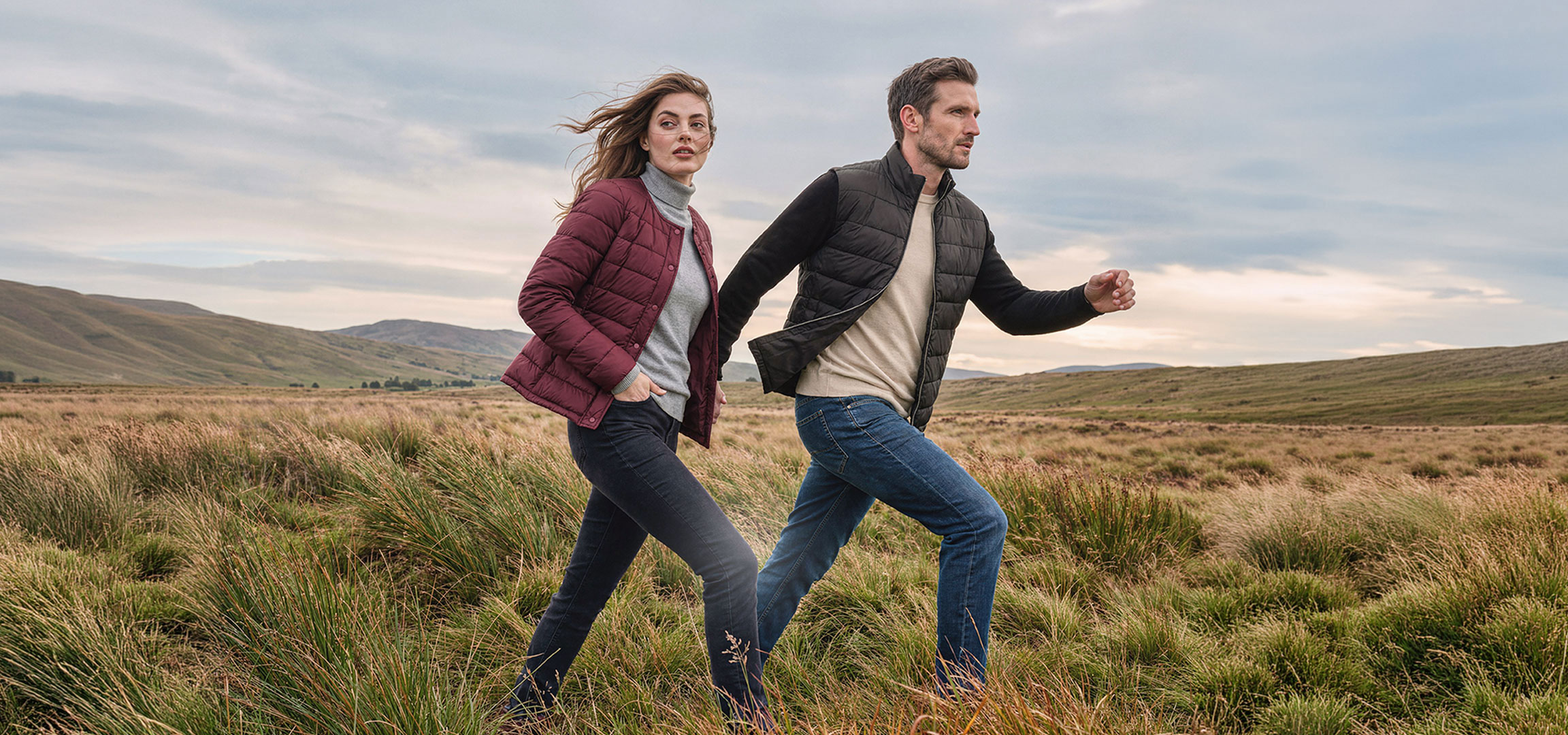 aiLabeling.aiLabelForImageAltText: Couple in lightweight jackets and jeans walks through a grassy, hilly landscape.