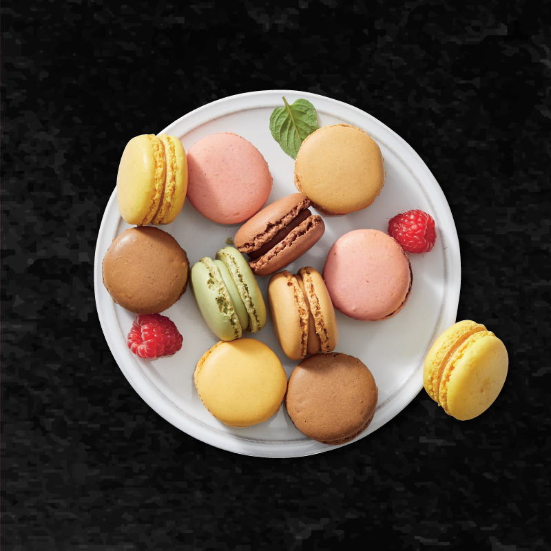 Assortment of colorful macarons on a white plate with raspberries and a mint leaf.