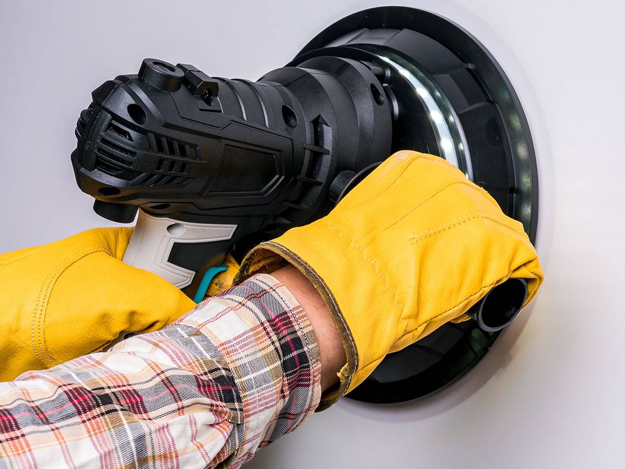 Electric wall sander with LED light, used by a person wearing gloves.