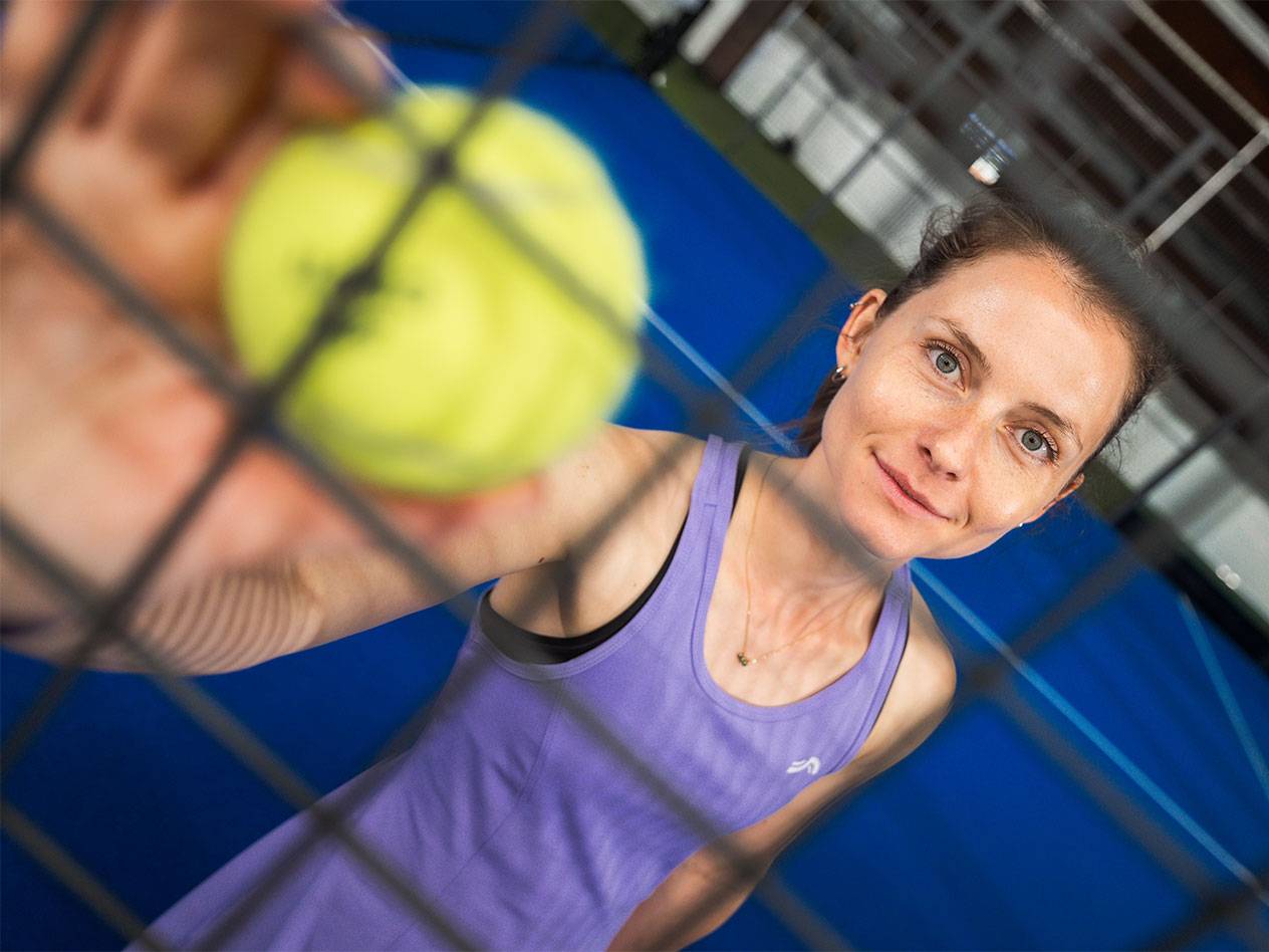 Woman on padel court holding yellow tennis ball through net.