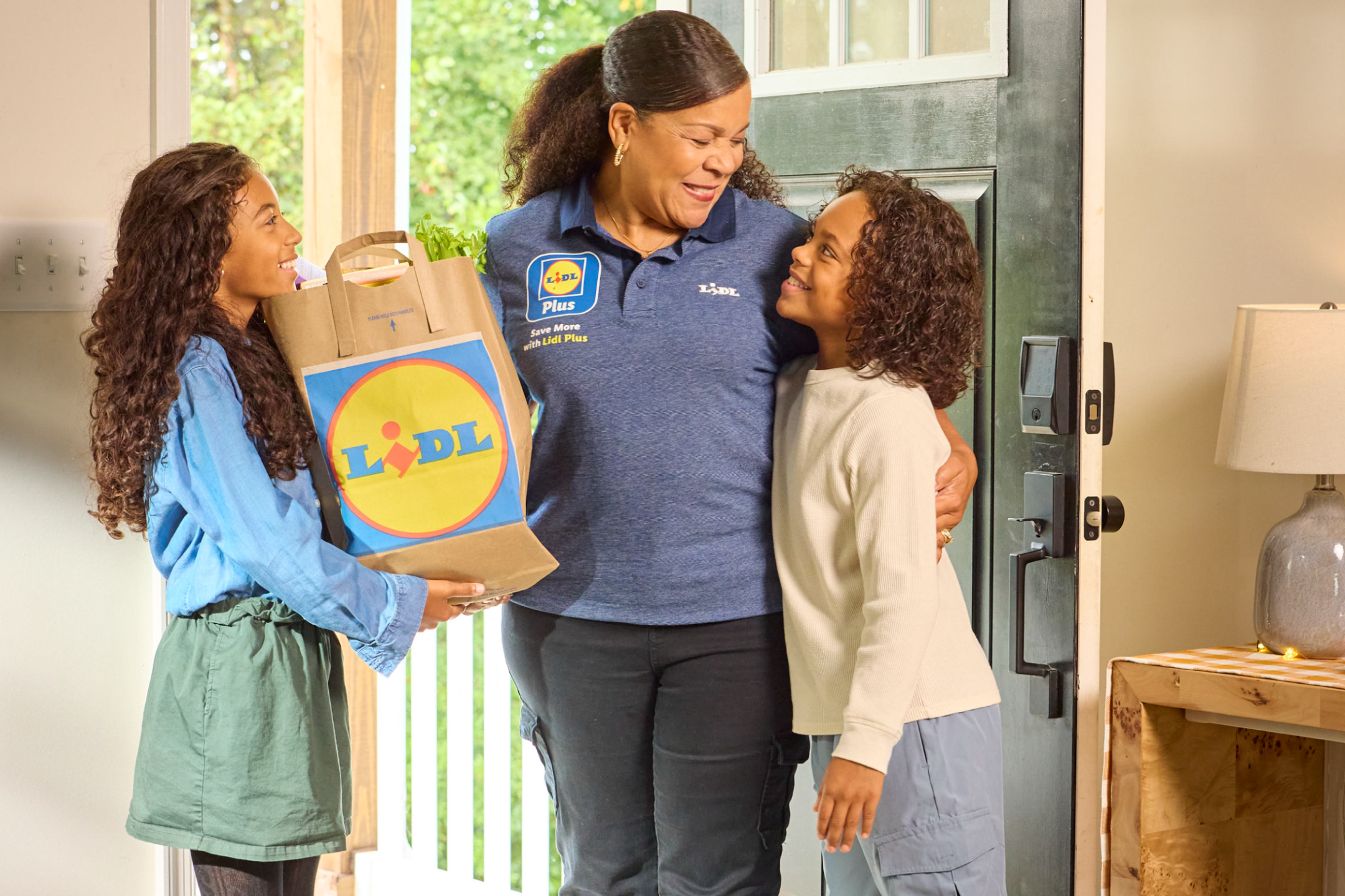 A delivery worker smiles as she delivers a paper bag of fresh groceries to two happy children.