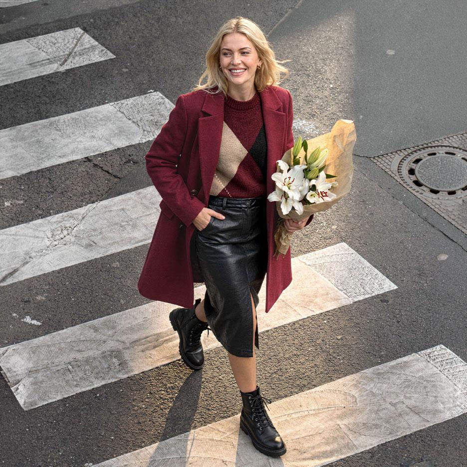 Smiling woman in a burgundy coat, argyle sweater, and leather skirt, crossing the street with a bouquet of lilies.