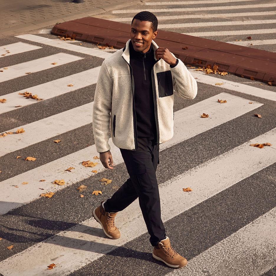 Smiling man in light fleece jacket, black shirt, and black cargo pants, walking on a crosswalk.