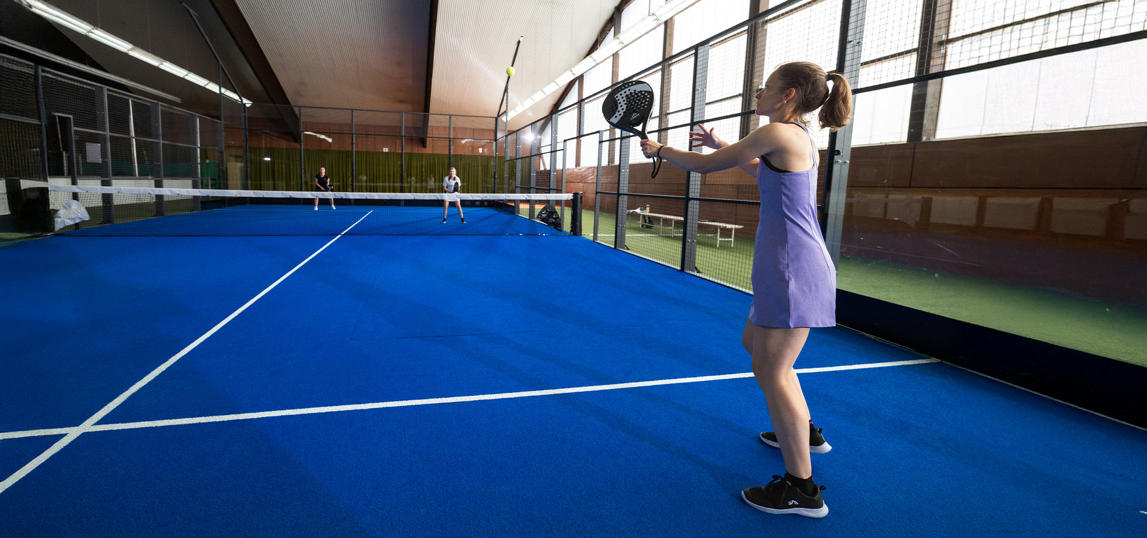 Lana Büttner plays padel on the court with a Crivit paddle, a yellow ball, and other players.