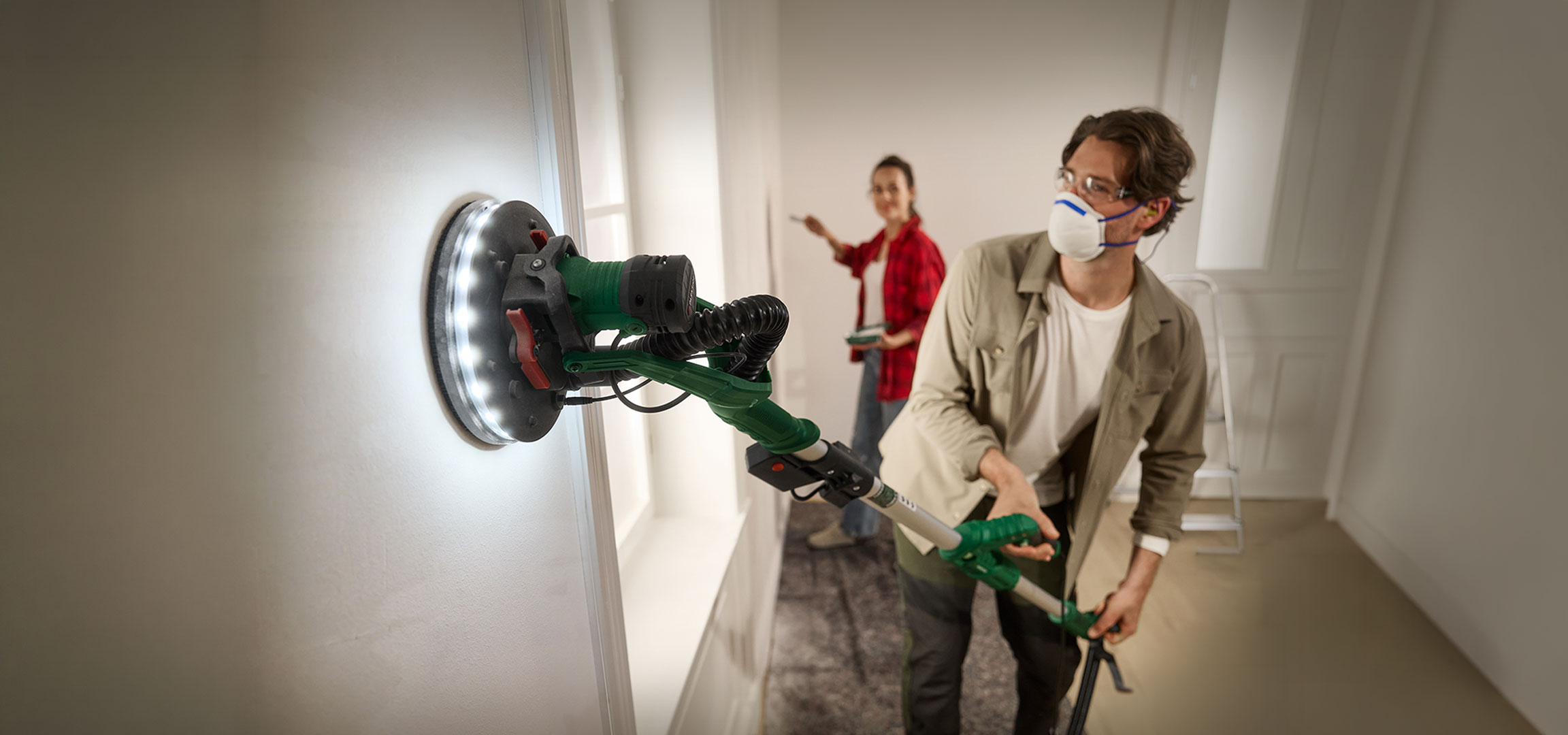 aiLabeling.aiLabelForImageAltText: Man sanding a wall with a long-reach sander, woman painting in the background.