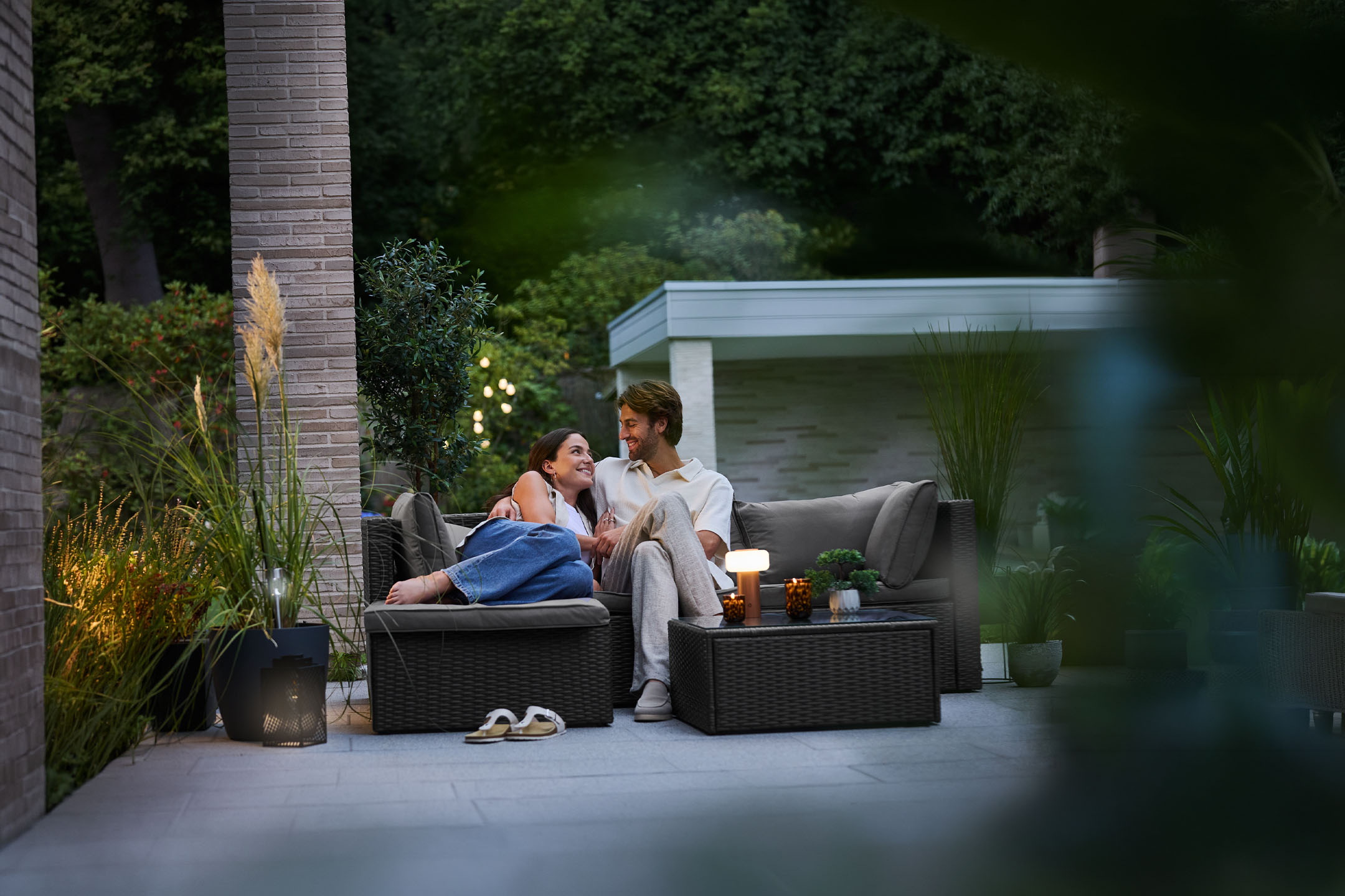 Couple relaxing on wicker garden furniture, illuminated by string lights and candles.