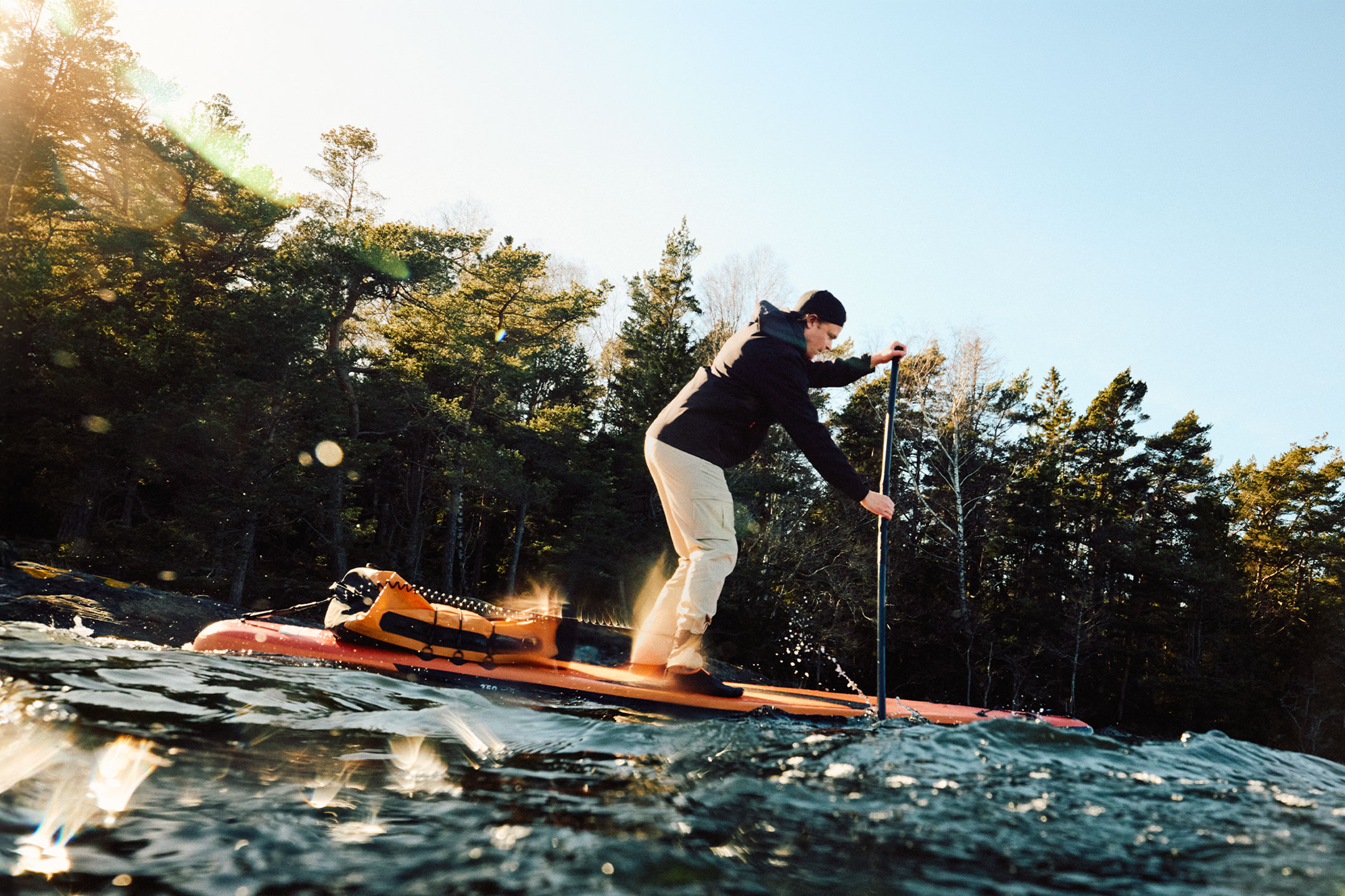 Man in beige trousers and black jacket paddleboarding on an orange paddleboard