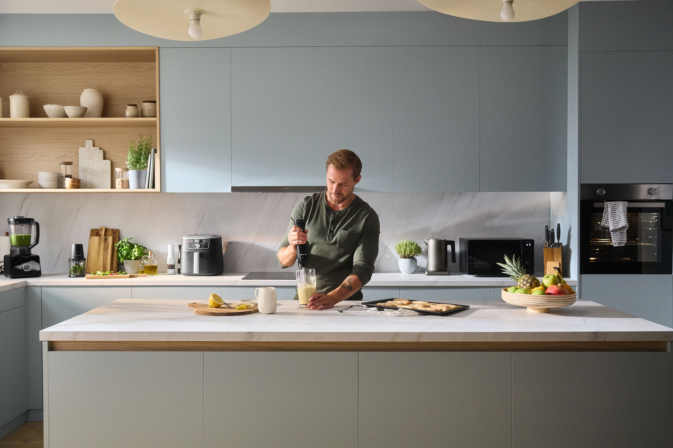 Man using an immersion blender in a modern kitchen with appliances and fruit.