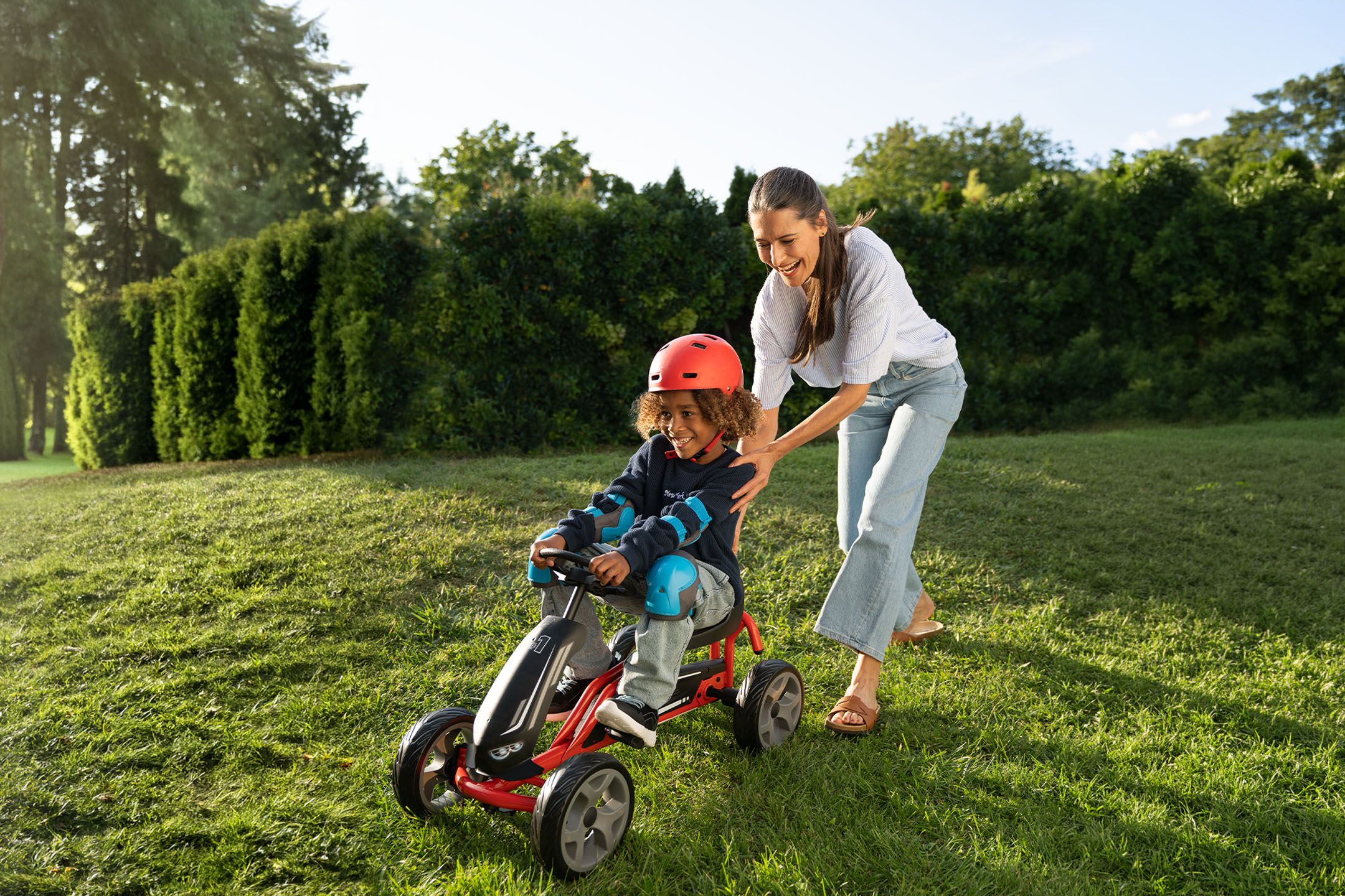 Mother and child playing outdoors with a pedal car, child wearing a helmet and pads.