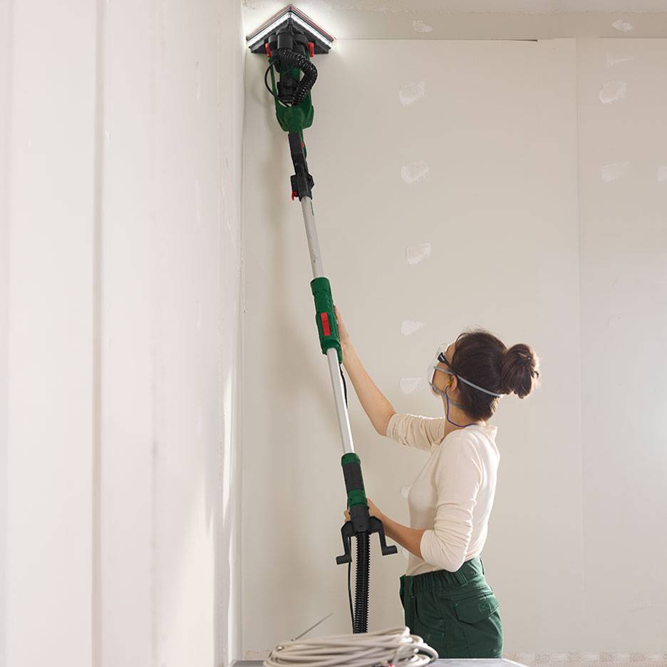 Woman sanding a wall with a telescopic wall sander in a corner.