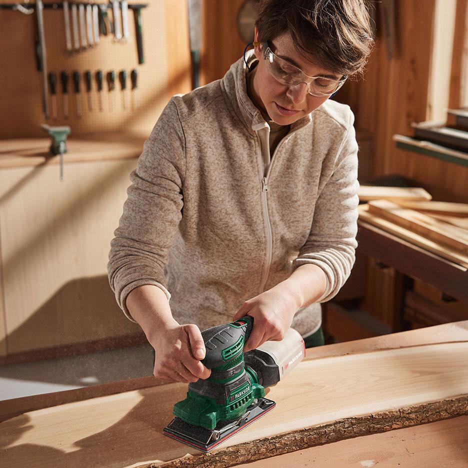 Woman with electric sander sanding a piece of wood in a workshop.