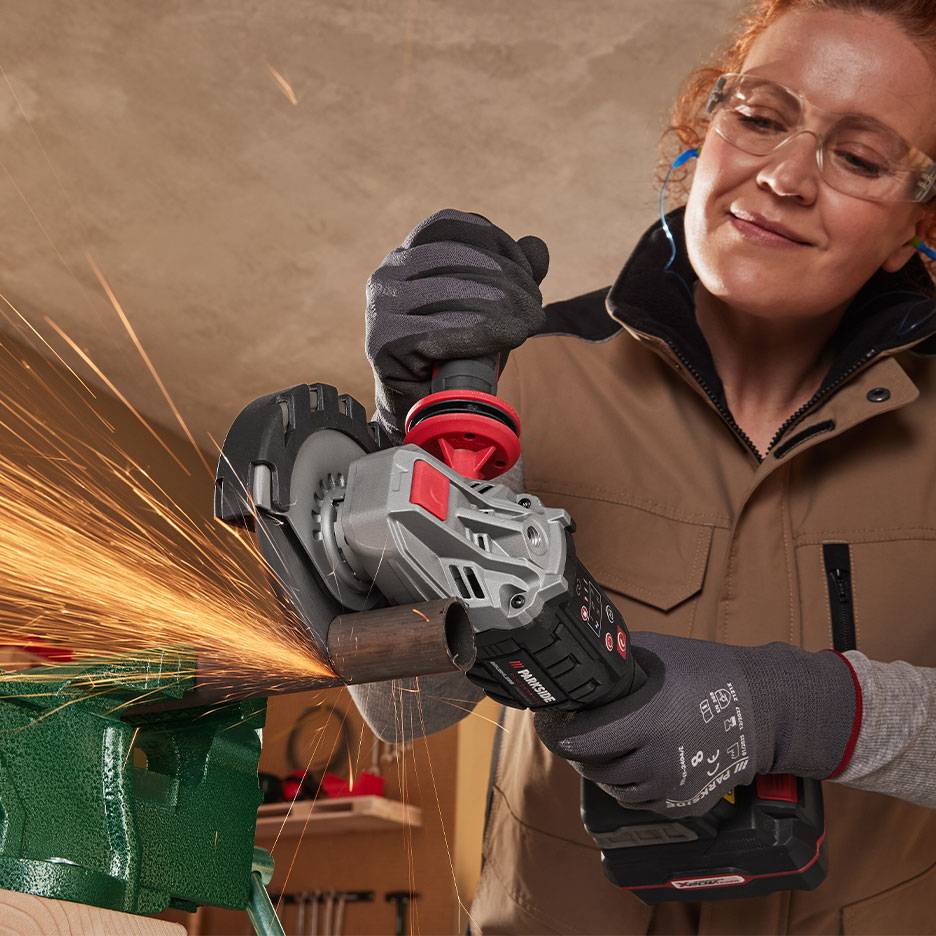 Woman in safety glasses using a cordless angle grinder to cut a metal pipe.