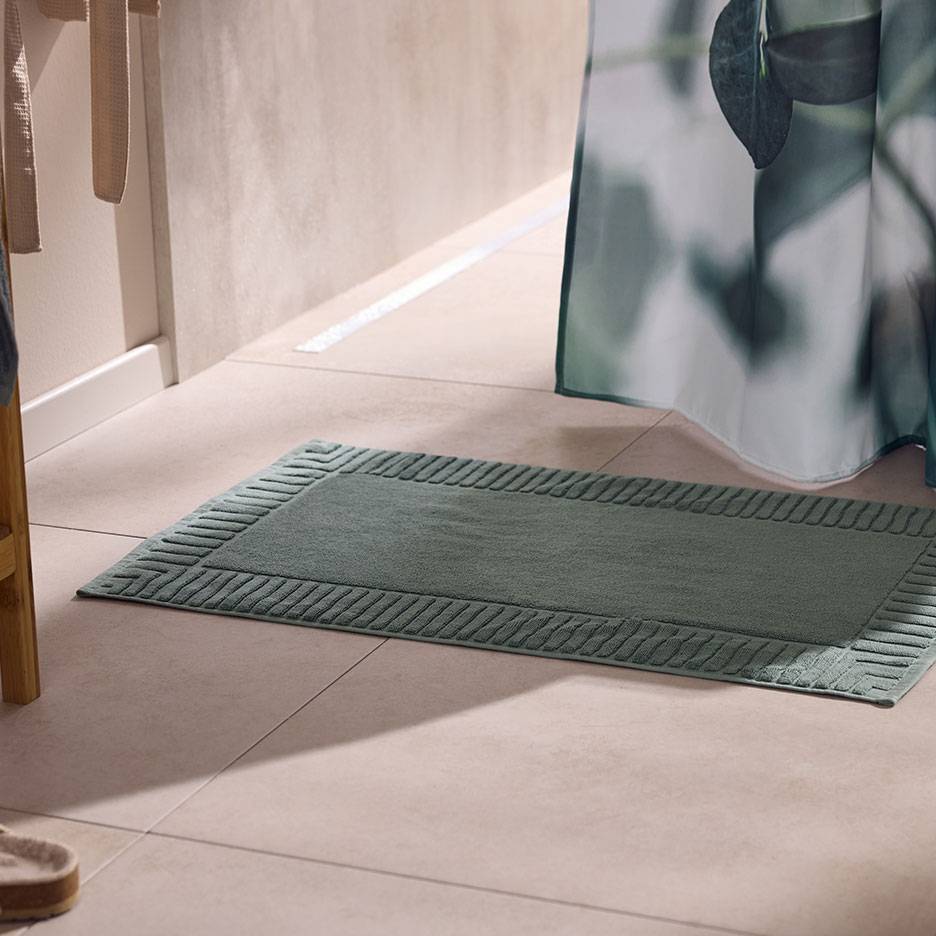 Green textured bath mat on light tiles, next to a shower curtain.