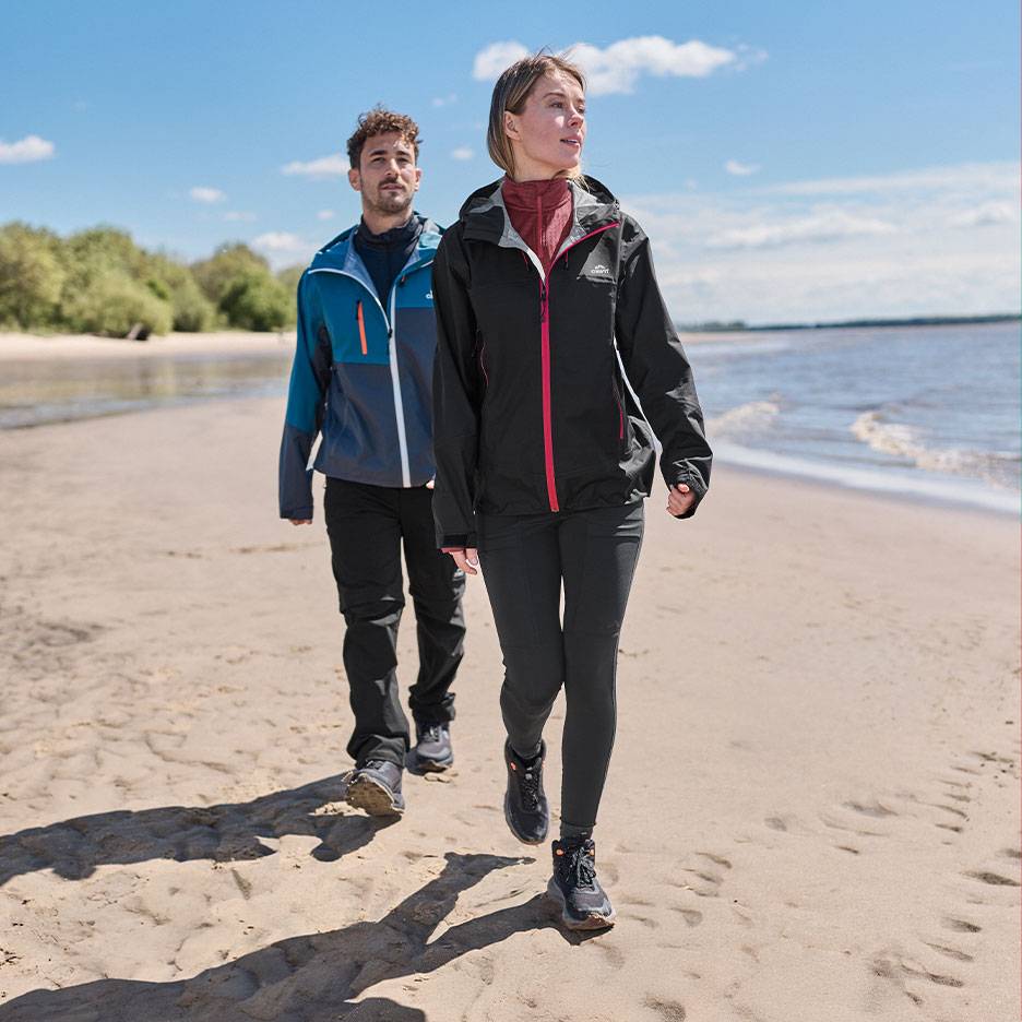 Couple in hiking jackets and trousers walking on a sandy beach by the sea.