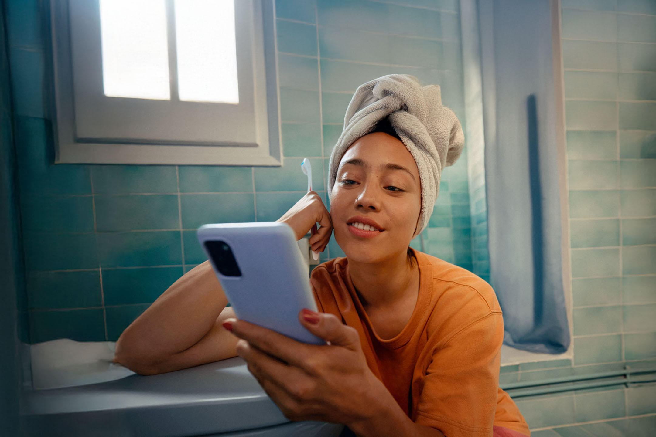 A woman with a towel on her head sitting in a bathtub using a smartphone.