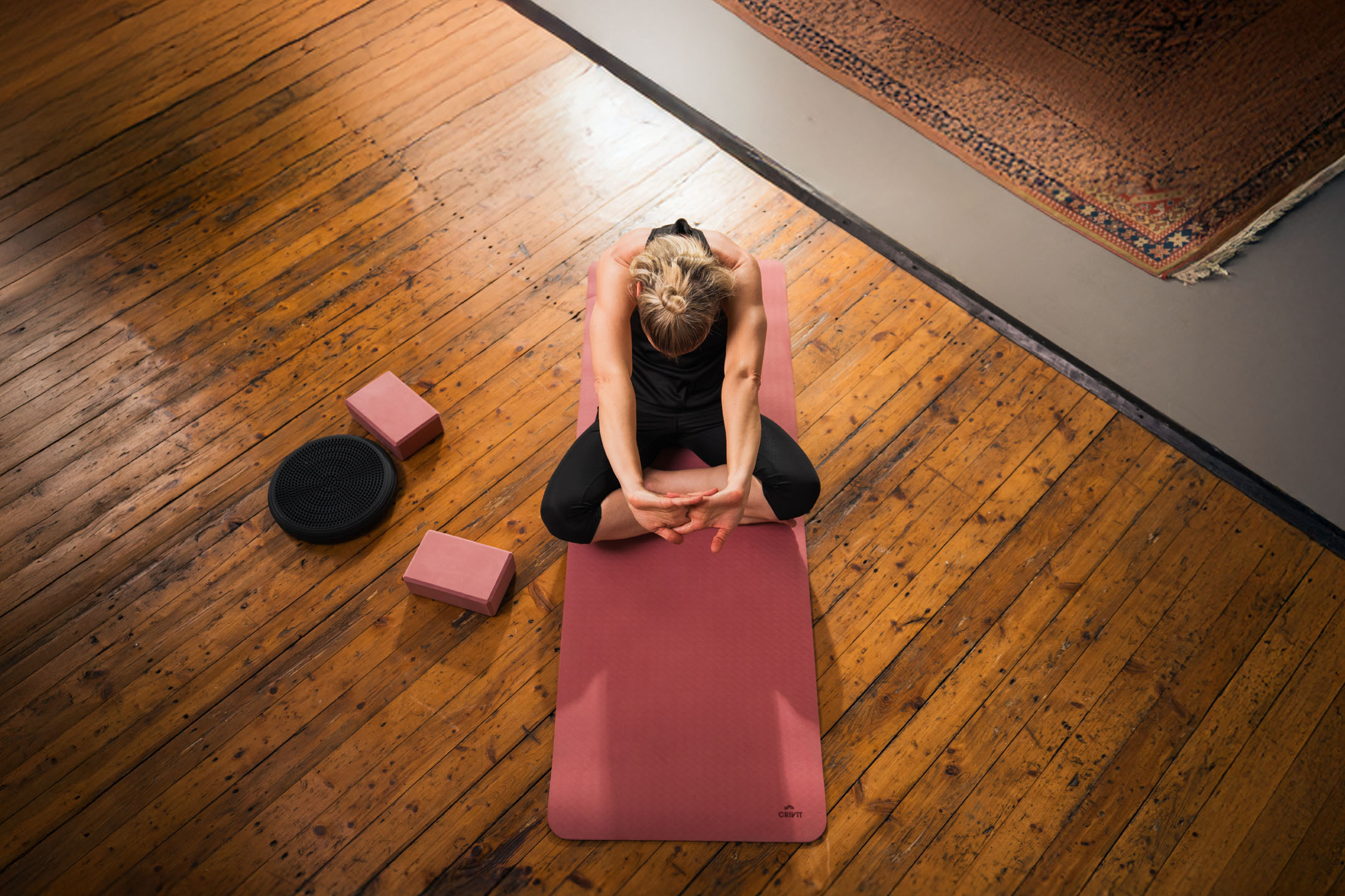 Mulher praticando yoga em um tapete rosa com blocos e disco de equilíbrio.