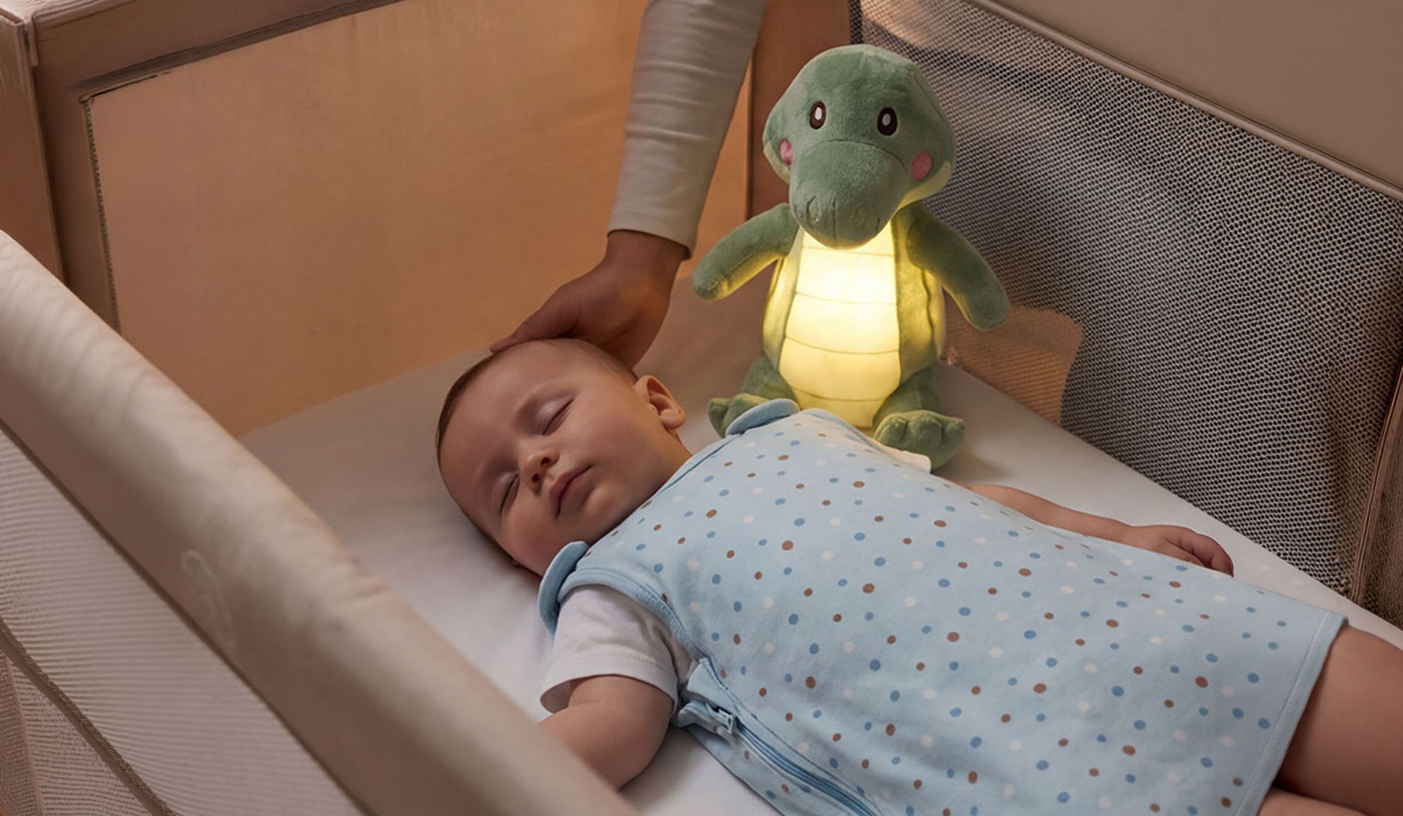 Baby sleeping in a cot with a soft toy and a parent's hand.