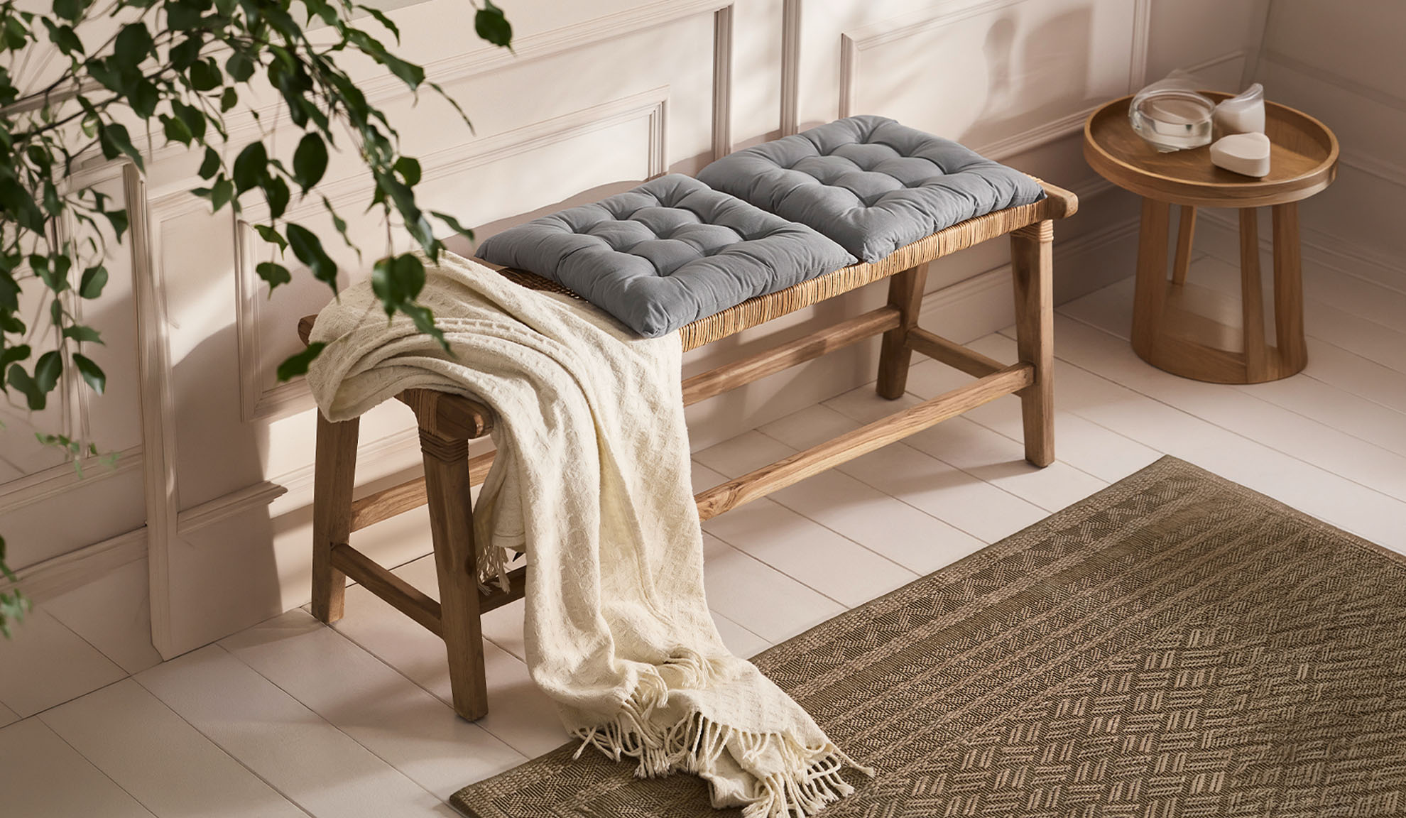Wooden bench with grey cushions and a throw, next to a wooden table with dishes, a rug on the floor.