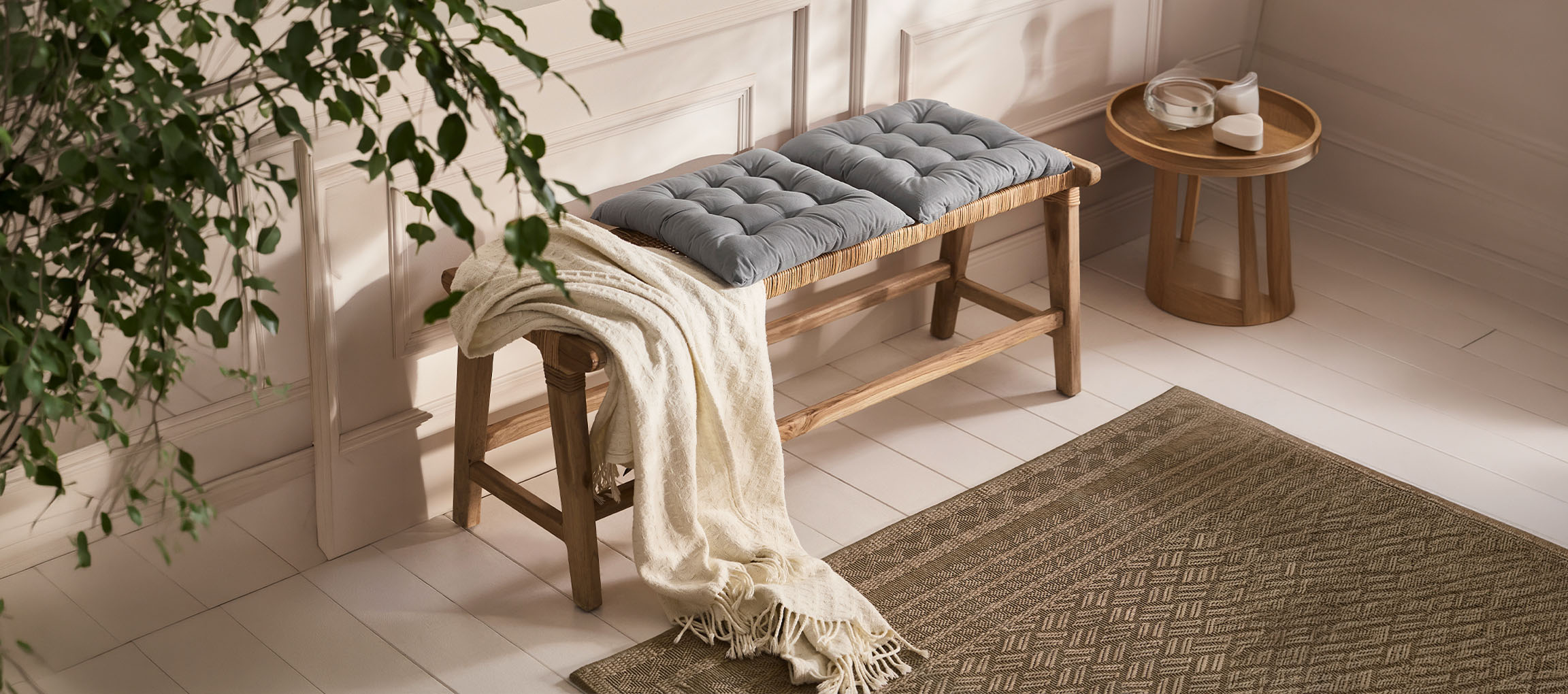 Wooden bench with grey cushions and a white throw on a light wooden floor with a rug.