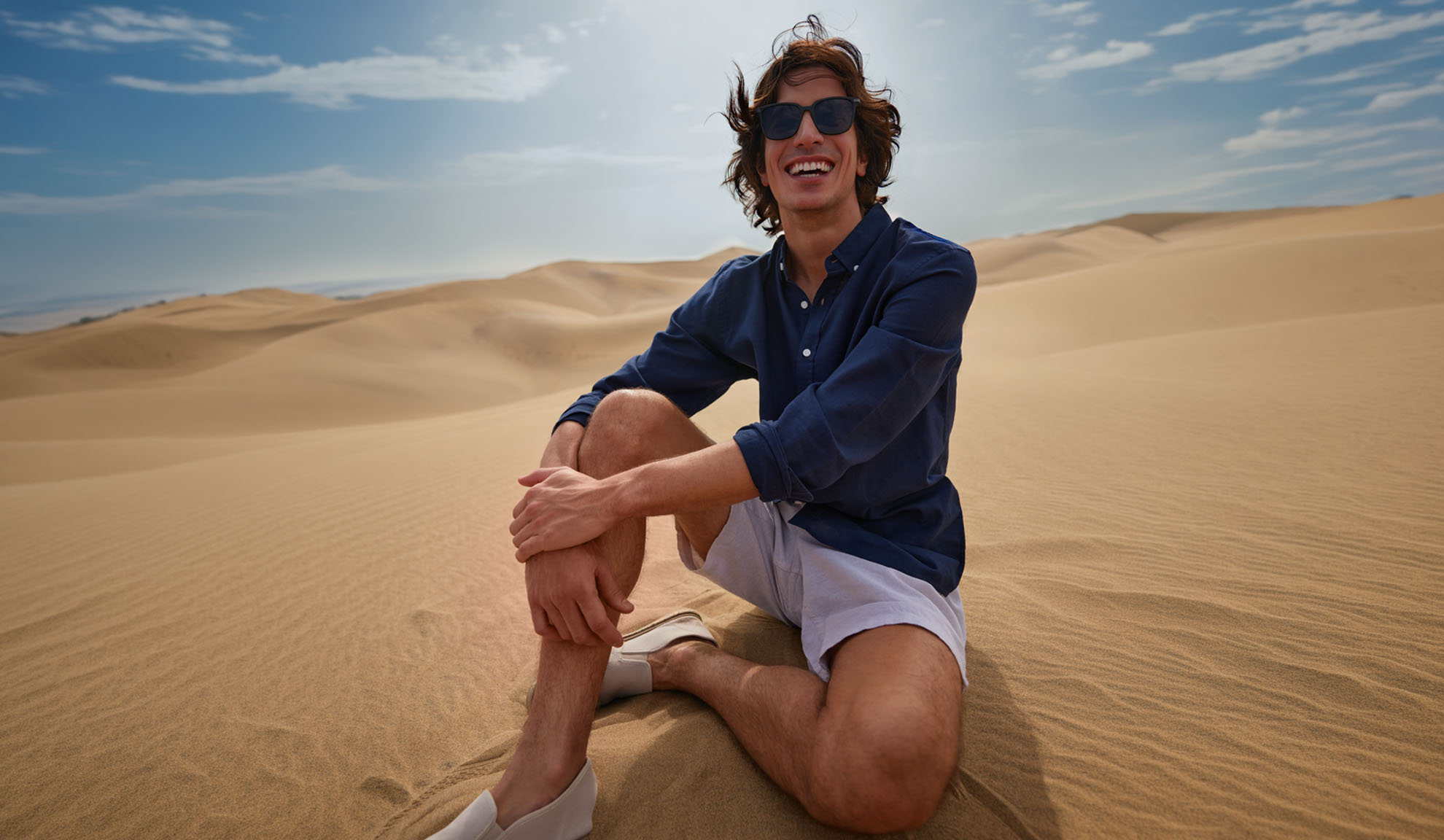 Man in dark blue shirt and light shorts sits on sand dunes, smiling.