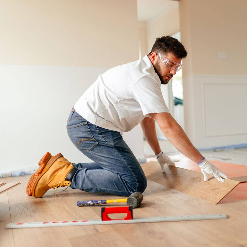 Man installing laminate flooring, wearing safety glasses and work boots.