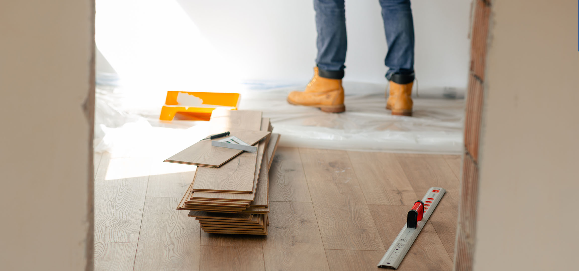 Laminate flooring planks and installation tools on a wooden floor, with a person in boots.
